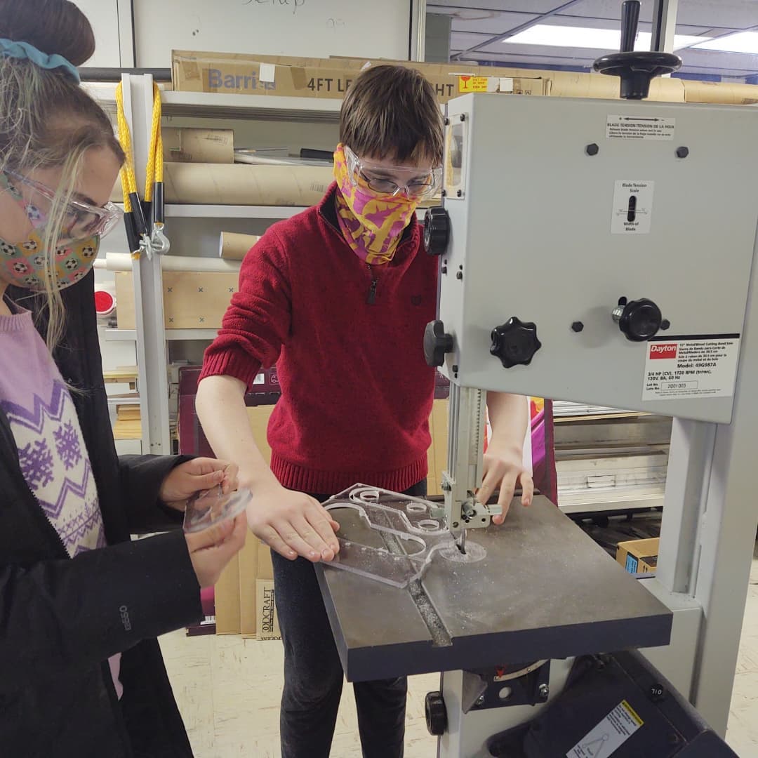 a student cutting a piece of plastic on a band saw while another student watches